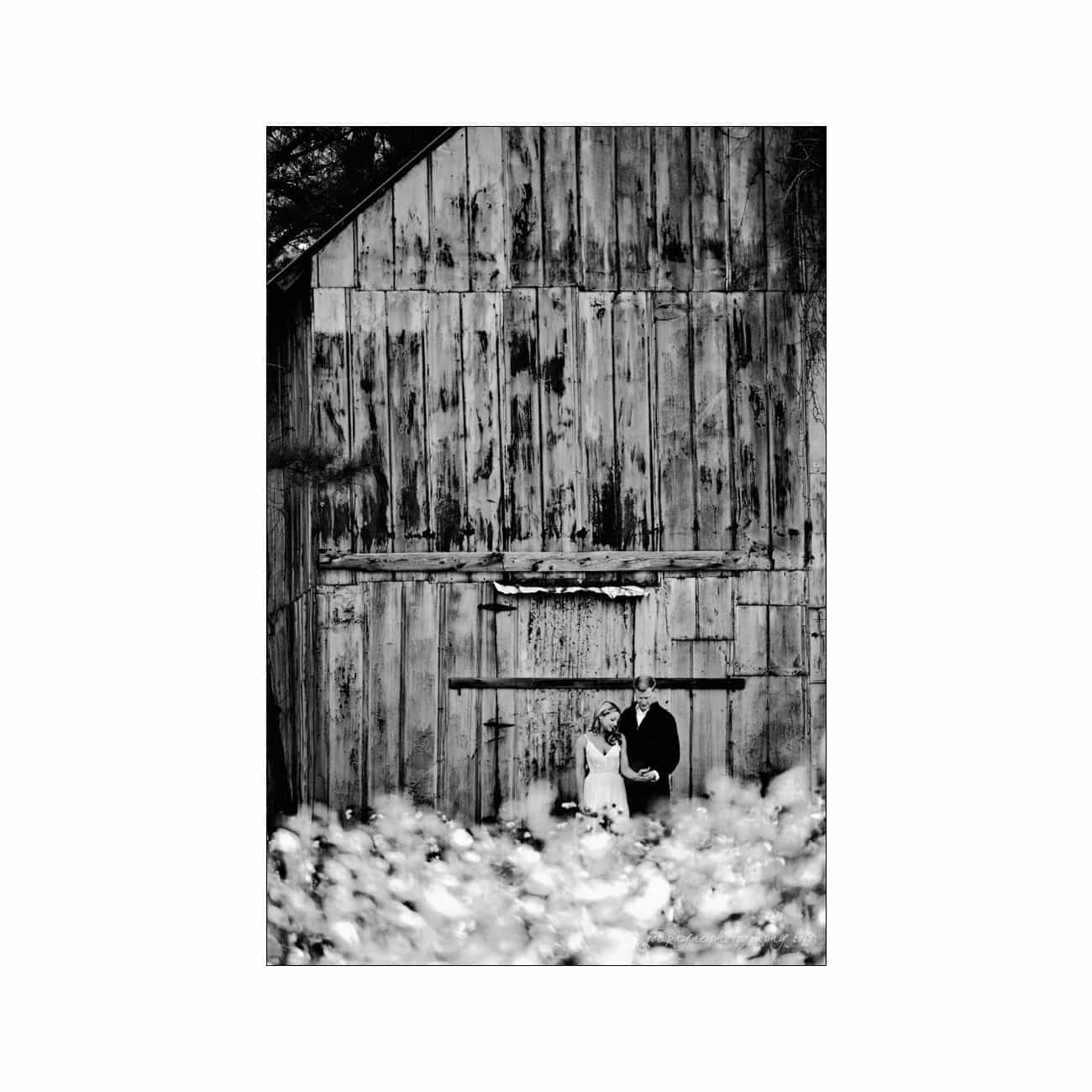 A black and white photo of a couple standing together in front of an old wooden barn. They are surrounded by blurry white flowers in the foreground, creating a soft, romantic atmosphere.