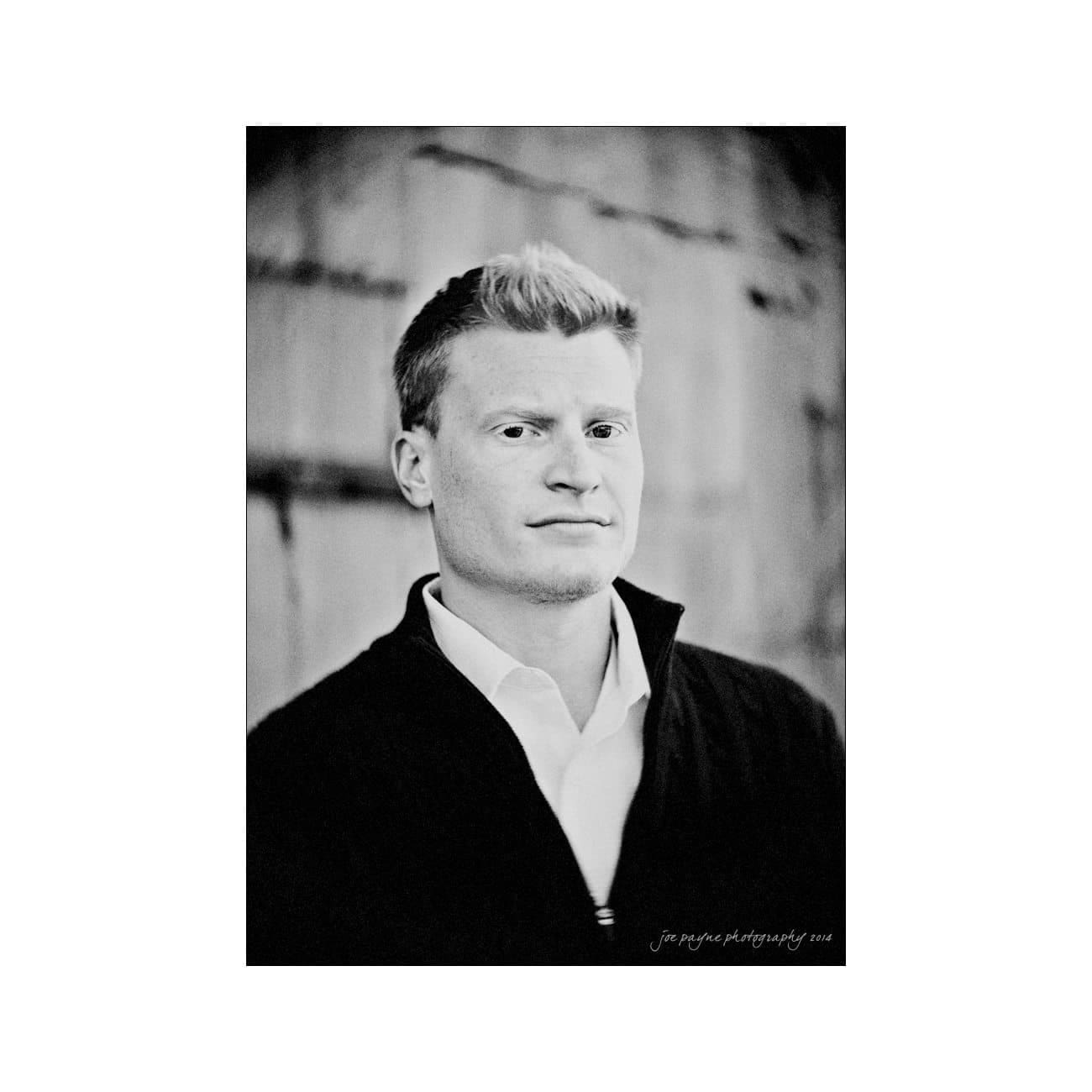 Black and white portrait of a person with short, light-colored hair, wearing a dark jacket over a white shirt, standing in front of a textured background.