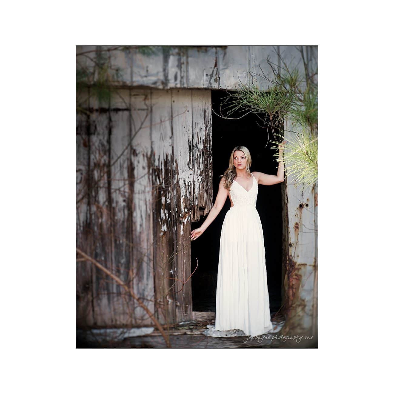 A woman in a flowing white dress stands in the doorway of an old, rustic wooden building, surrounded by greenery. The door is partially open, and she gazes thoughtfully into the distance.