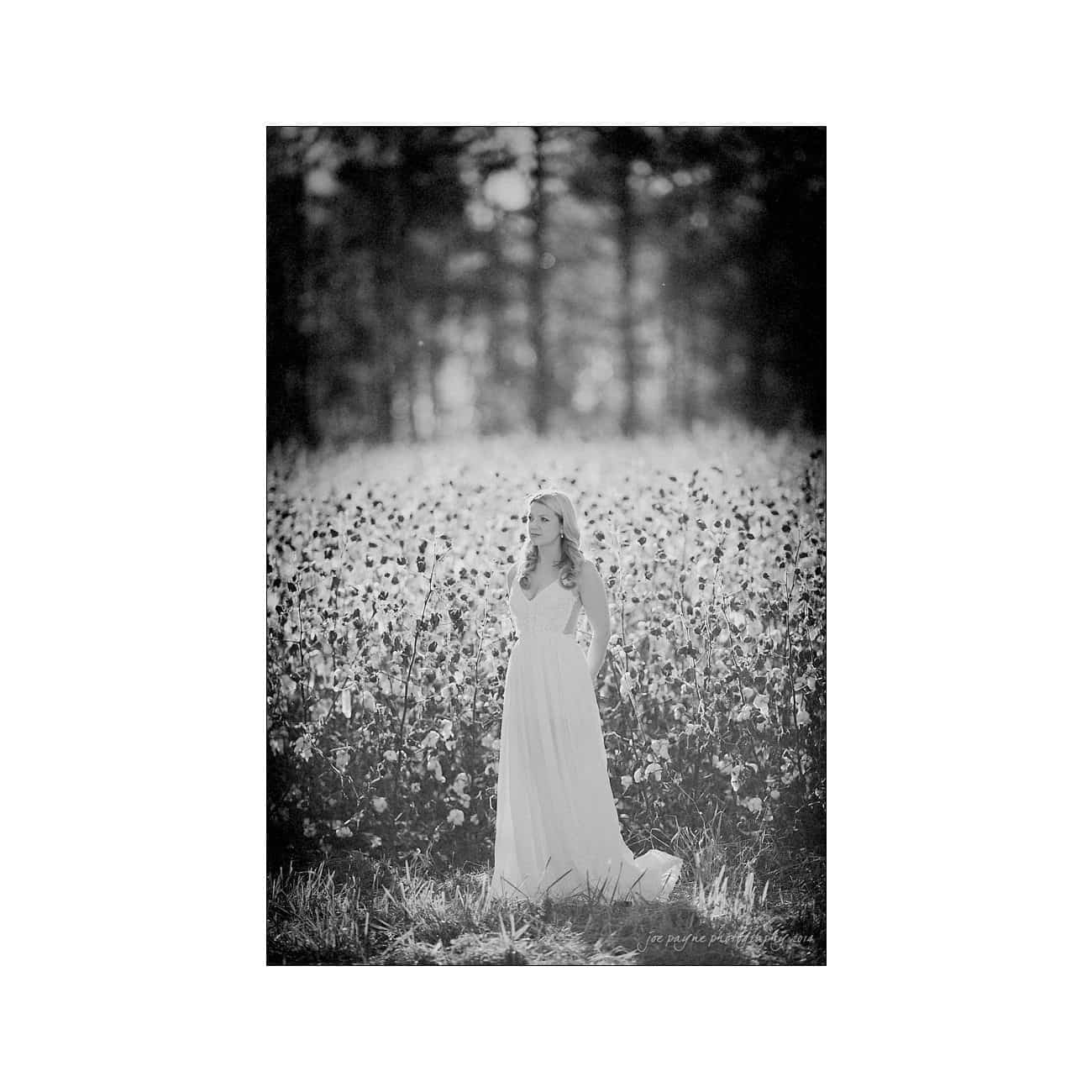 A woman in a long, flowing white dress stands amidst a field of flowers. The image is in black and white, with a blurred forest background, giving a serene and timeless feel.