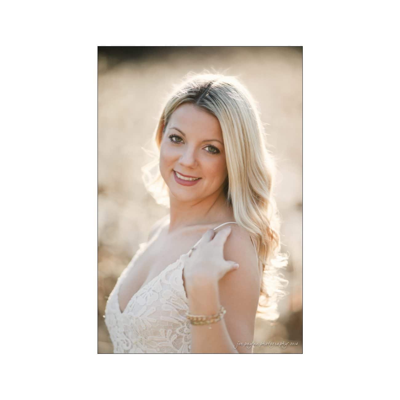A woman with long blonde hair smiles softly at the camera. She is wearing a white lace dress and touches her shoulder, standing against a softly blurred background with warm, natural lighting.