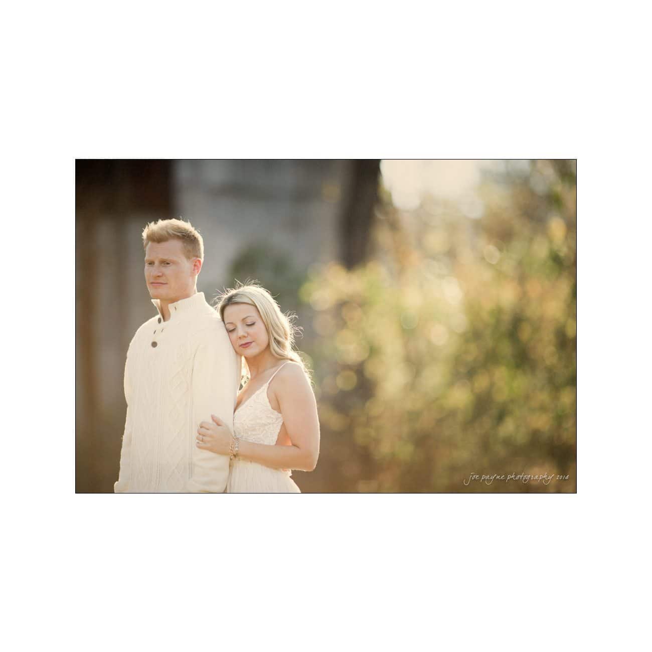 A couple stands outdoors in soft, golden sunlight. The man wears a cream sweater and looks ahead, while the woman, in a delicate white dress, gently leans on his shoulder with her eyes closed, creating a serene and intimate moment.