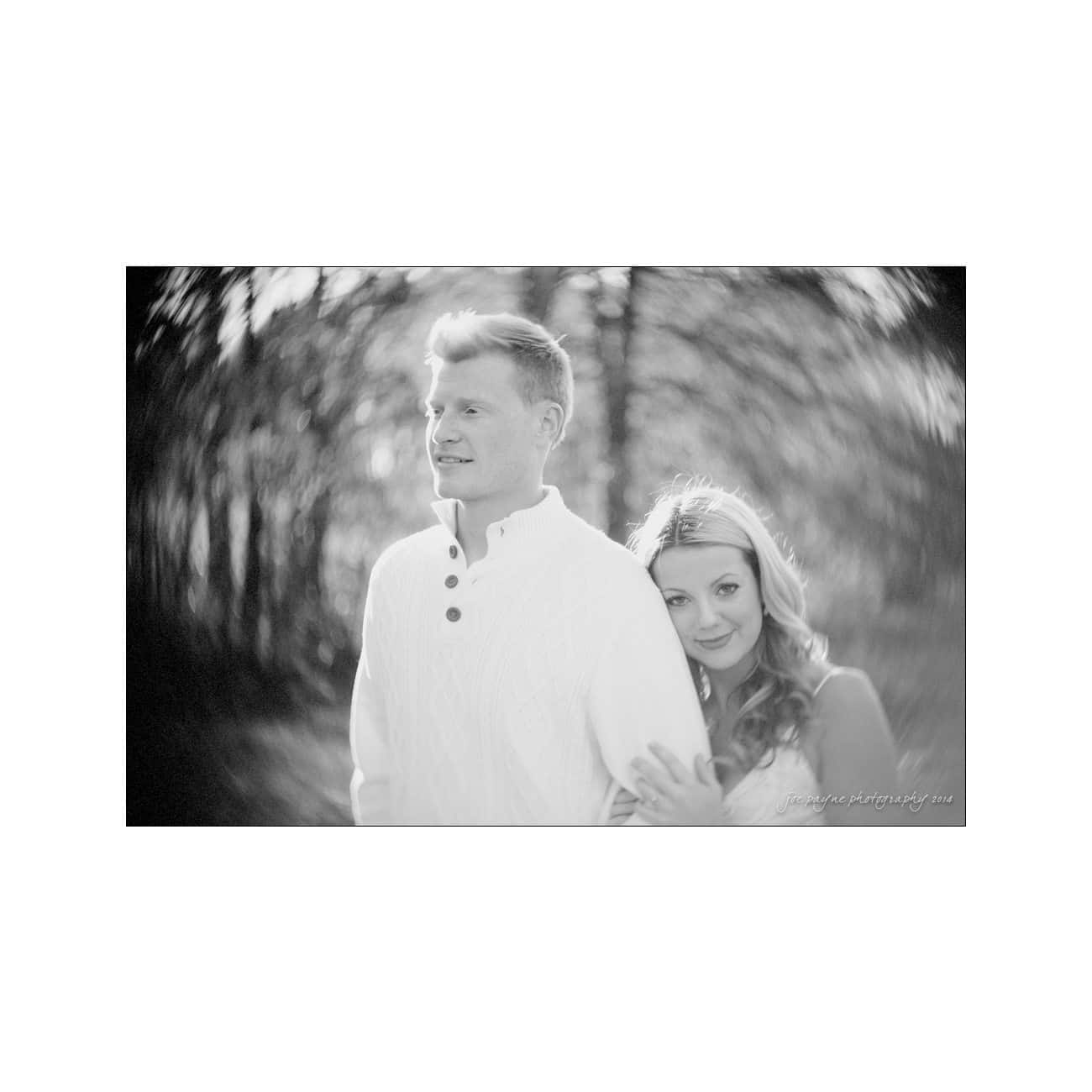 A black and white photo of a couple outdoors. A woman with long hair rests her head on a mans shoulder. Both are smiling slightly, with the background showing blurred trees and light effects creating a dreamy atmosphere.