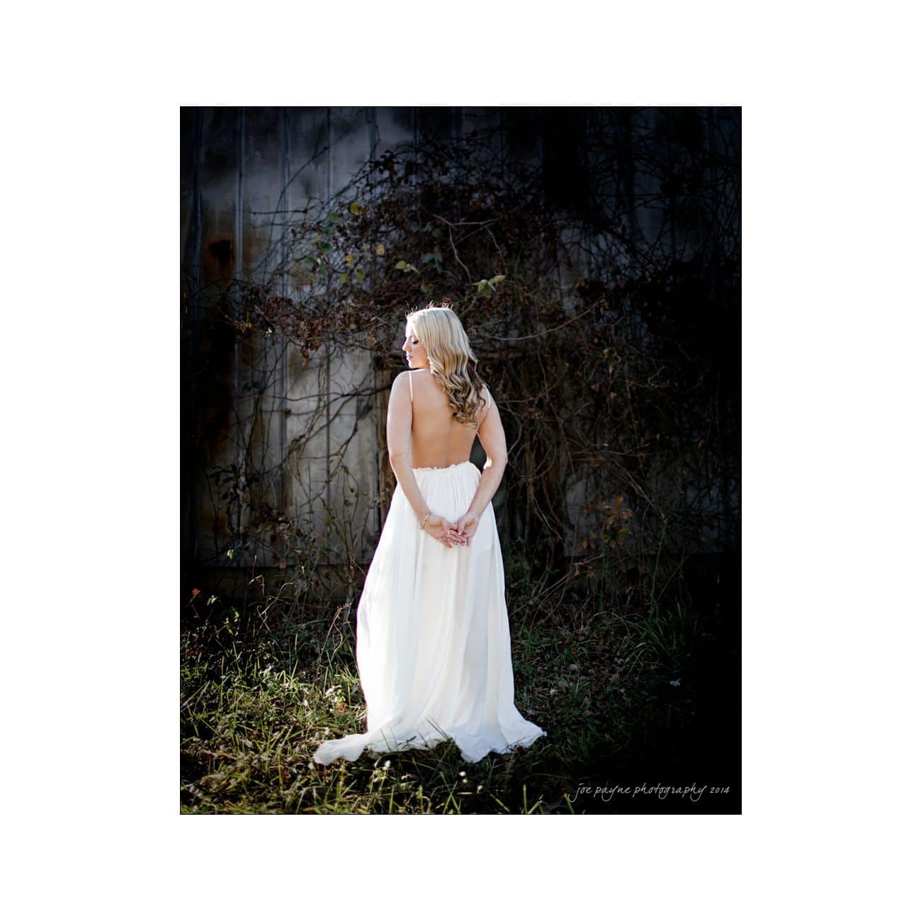 A woman in a backless, flowing white dress stands outdoors against a rustic wooden wall entwined with vines. Her hands are clasped behind her back as she gazes away from the camera. Sunlight highlights her blonde hair, creating a serene scene.