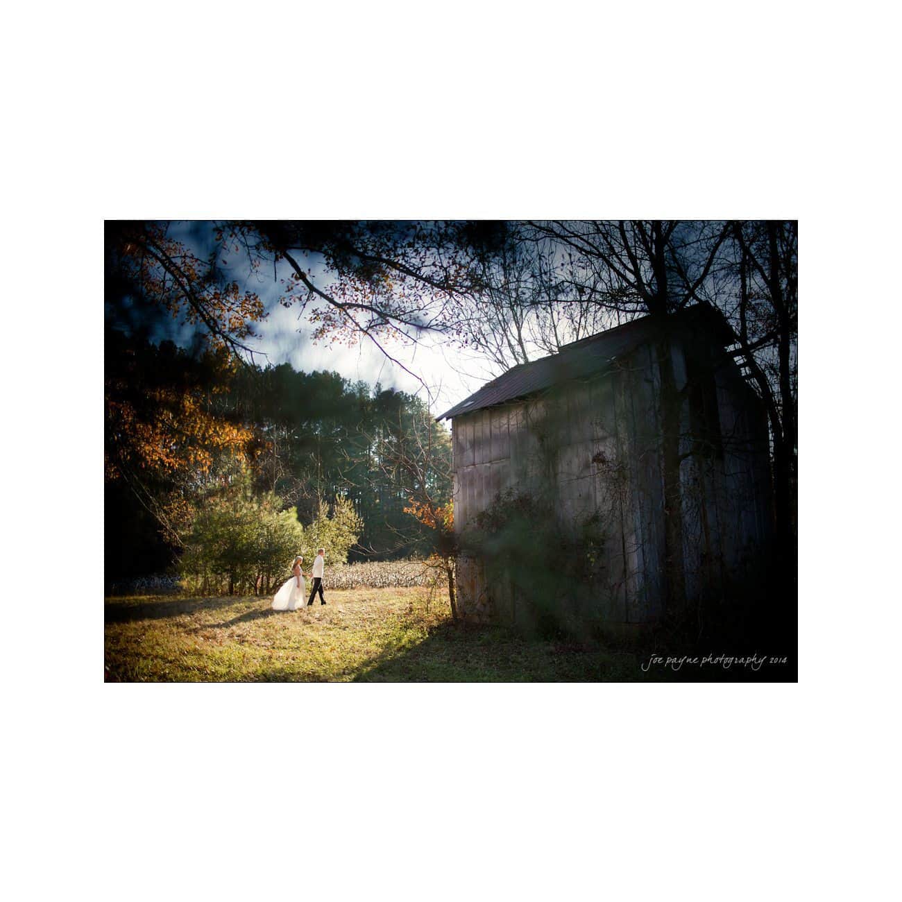 A couple walks hand in hand in a sunlit field, surrounded by trees with autumn foliage. An old barn stands to the right. The sky is partly cloudy, creating a serene and romantic atmosphere.