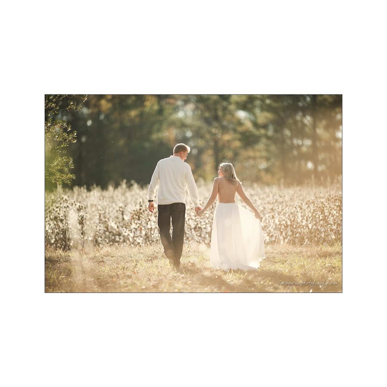 A bride and groom walk hand in hand through a sunlit field. The bride wears a long white dress, and the groom is in a white shirt and dark pants. The scene is serene with a soft, golden glow from the sunlight filtering through the trees.