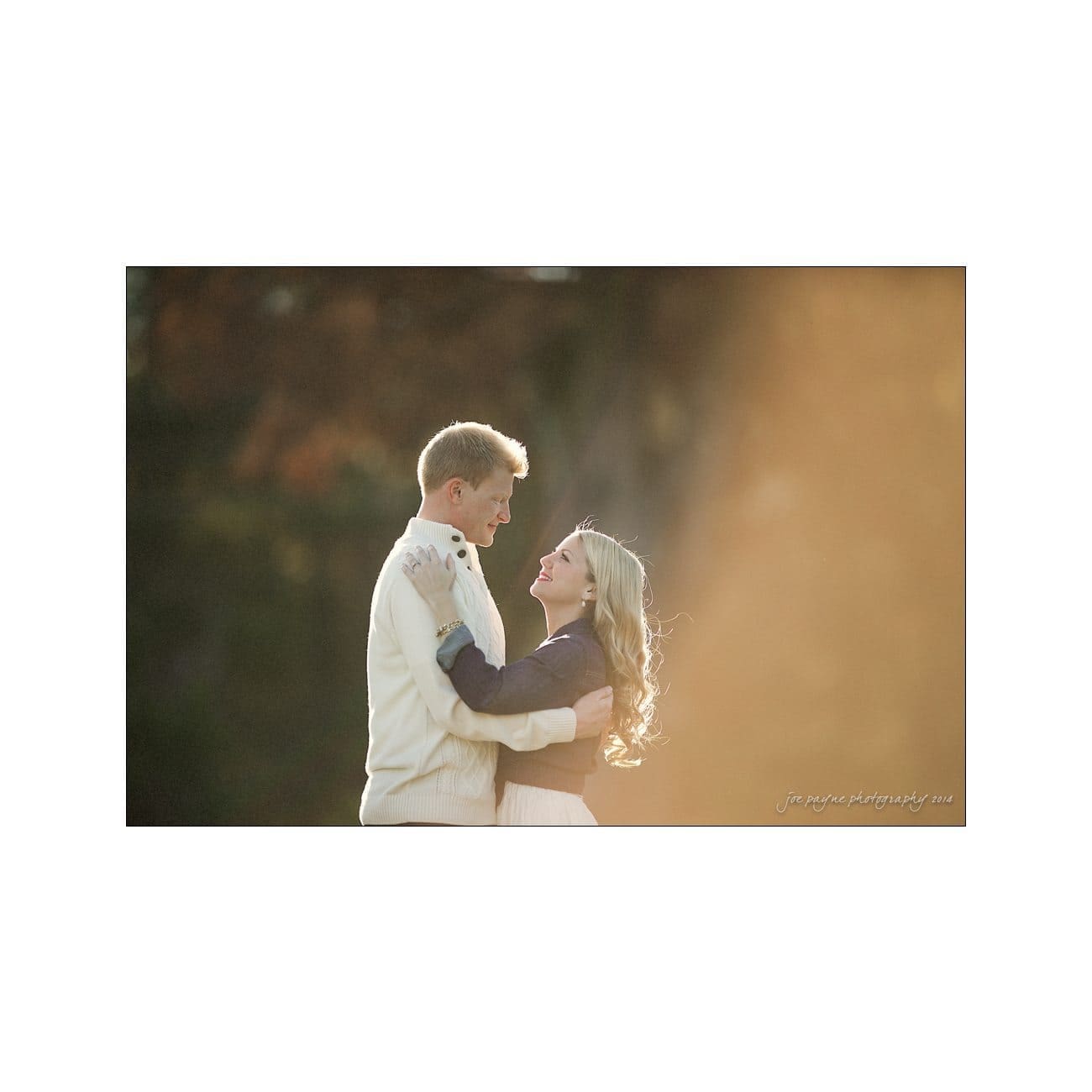 A couple stands outdoors in warm sunlight, dressed in cozy sweaters. The man holds the woman as they gaze into each others eyes, smiling, with a blurred natural background behind them.
