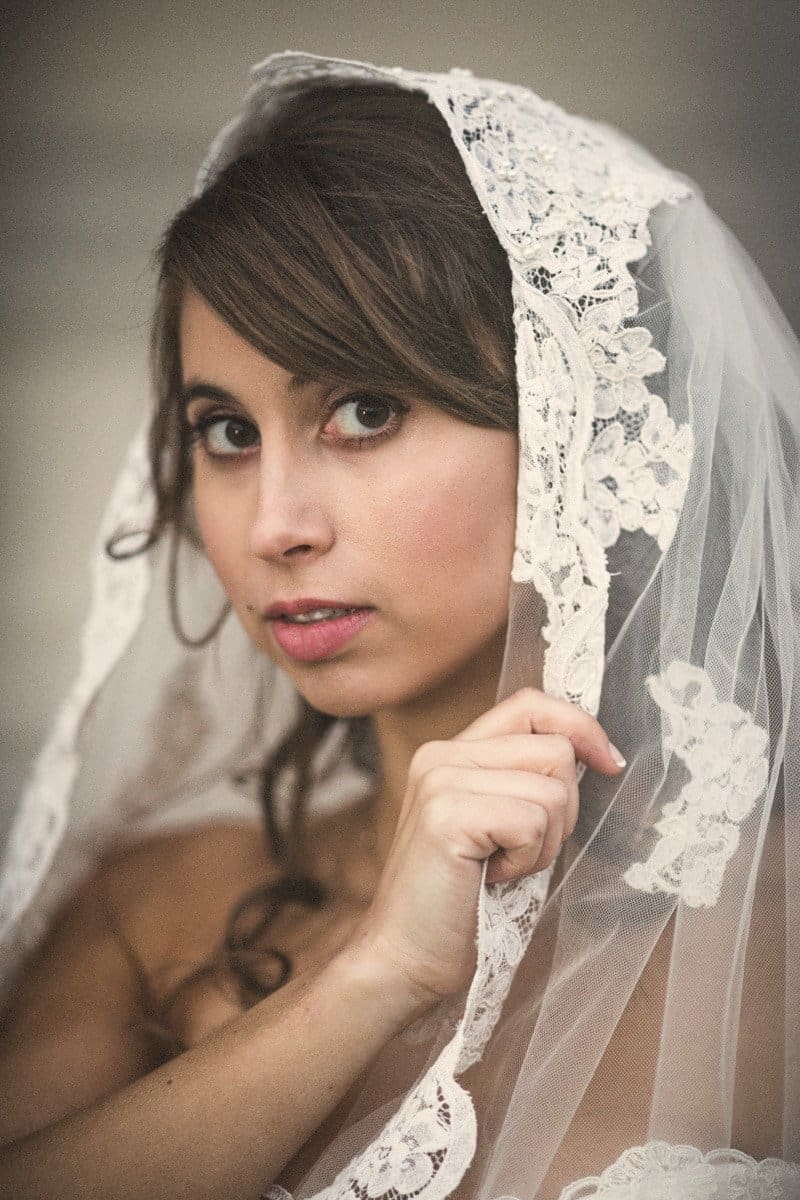 A bride with long brown hair gazes softly while holding a lace veil over her head. The lace features floral patterns, and her expression is calm and serene against a neutral background.