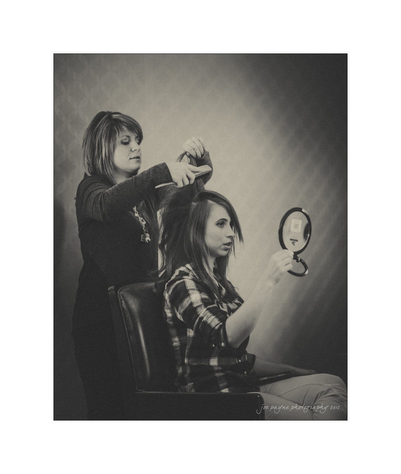A hairstylist works on a seated womans hair, who holds a small mirror in front of her. The setting has a vintage or classic ambiance, captured in black and white.