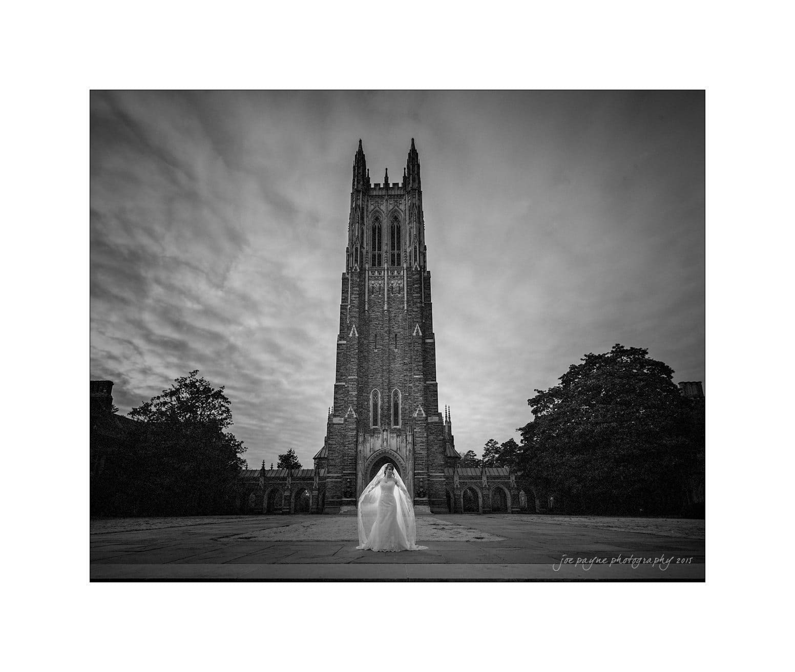 A black and white photo of a Gothic-style cathedral tower with a person in a long garment standing in the foreground. The sky is overcast, and trees line the path leading to the cathedral.