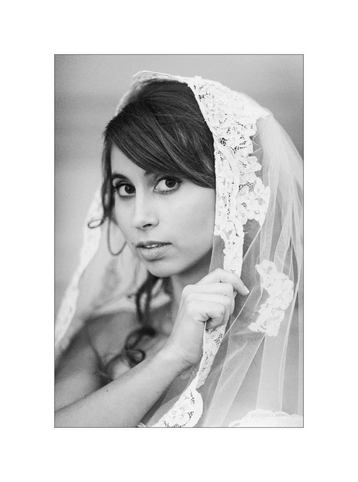 A woman gazes at the camera, partially covered by a lace veil. Her expression is thoughtful and serious. The black and white photo highlights the delicate texture of the veil and her soft features.