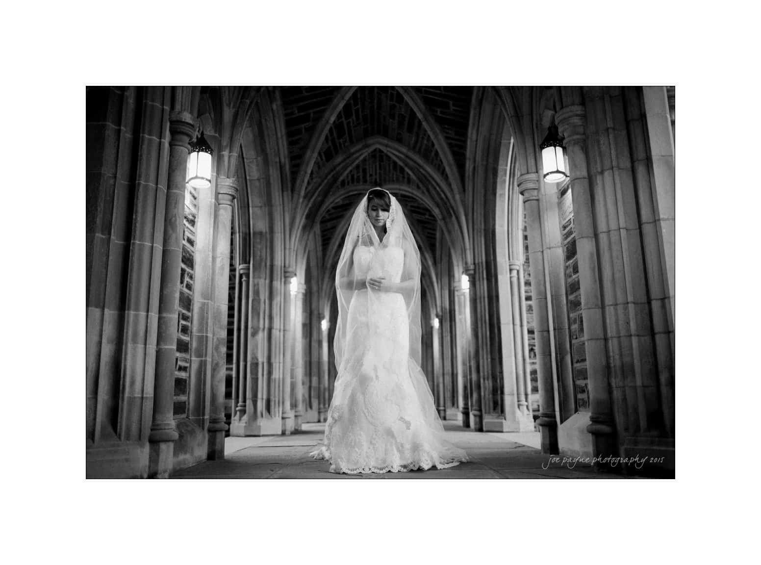 A bride in a lace dress and veil stands elegantly in a dimly lit, arched stone corridor. The scene is in black and white, highlighting the intricate architecture and the brides serene expression.