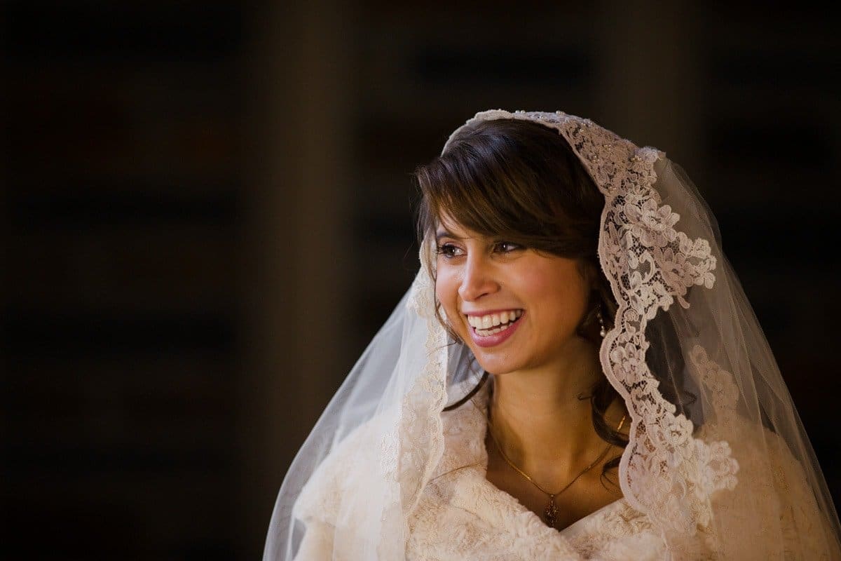 A bride smiles warmly while wearing a white, lace-trimmed veil and gown. Her dark hair is styled in soft waves. The background is dimly lit, highlighting her joyful expression.