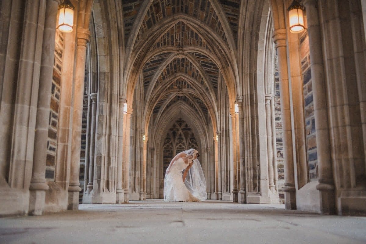 A bride in a white gown and veil stands under a series of dramatic stone archways, lit by warm lanterns. The architectural setting creates a serene and elegant atmosphere, with intricate details in the ceiling and walls.