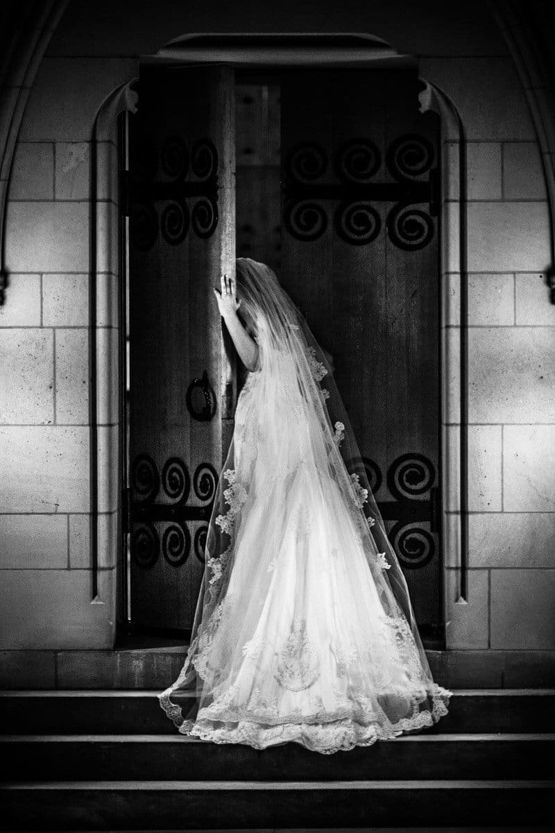 A bride in a long, lace-embroidered gown and veil stands at a large wooden door with intricate wrought iron designs. She rests one hand on the door, partially ajar, while standing on stone steps, captured in black and white.