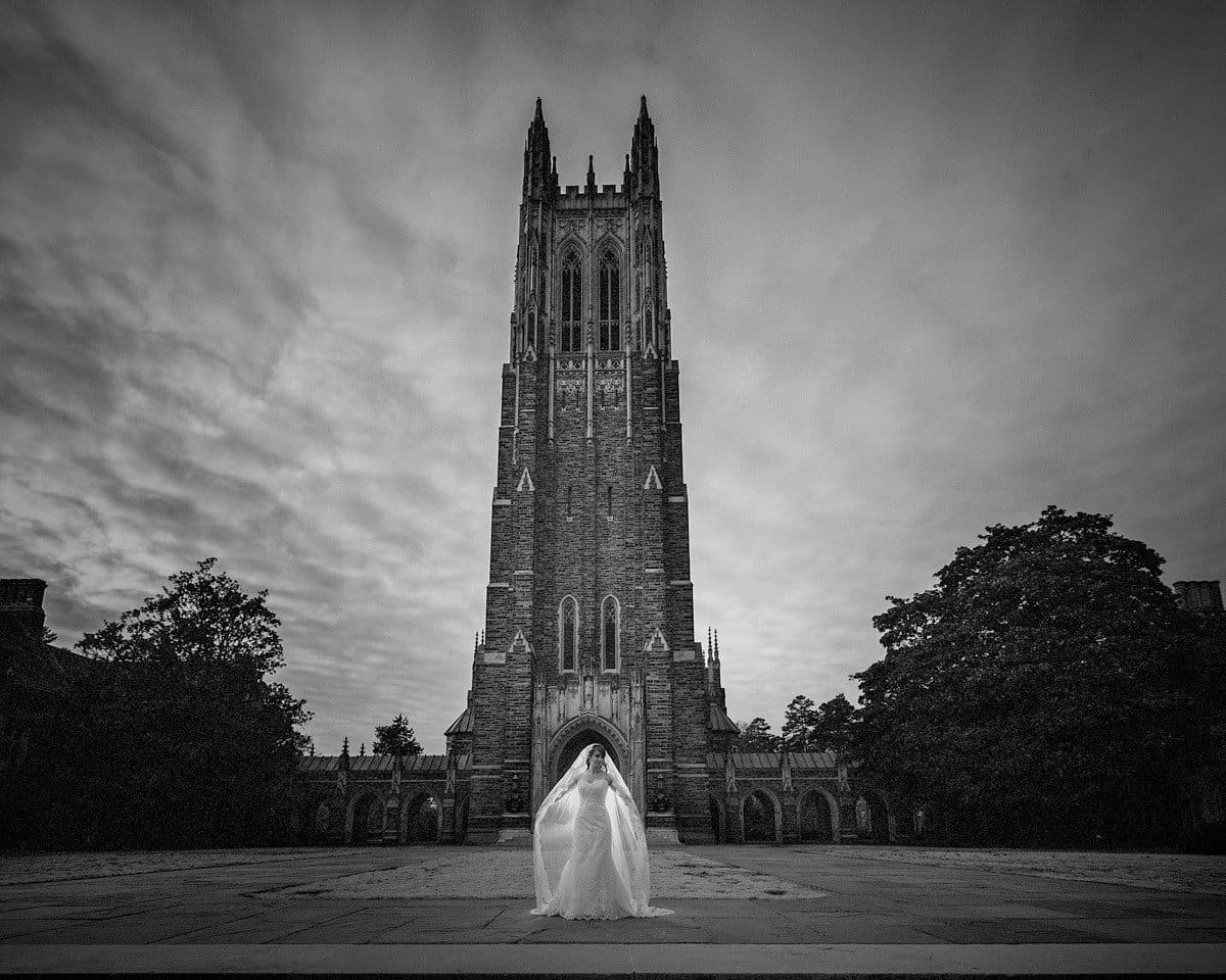 A bride and groom stand in front of a tall, Gothic-style cathedral tower under a dramatic sky. The black and white photo emphasizes the architectural details and the couples elegant presence in the foreground.
