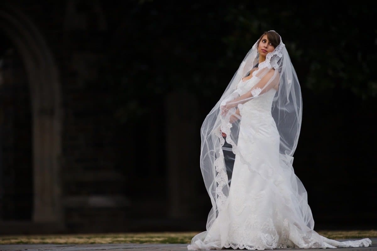 A bride in a white wedding dress with a long veil stands outdoors, with dark foliage in the background. She holds part of the veil in her hand and gazes upward.