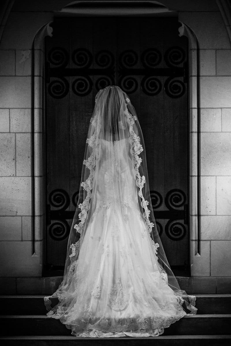 A bride in a long, flowing veil and gown stands with her back to the camera, facing a large, ornate wooden door with spiral designs. The setting has dim lighting, creating a dramatic and elegant atmosphere.
