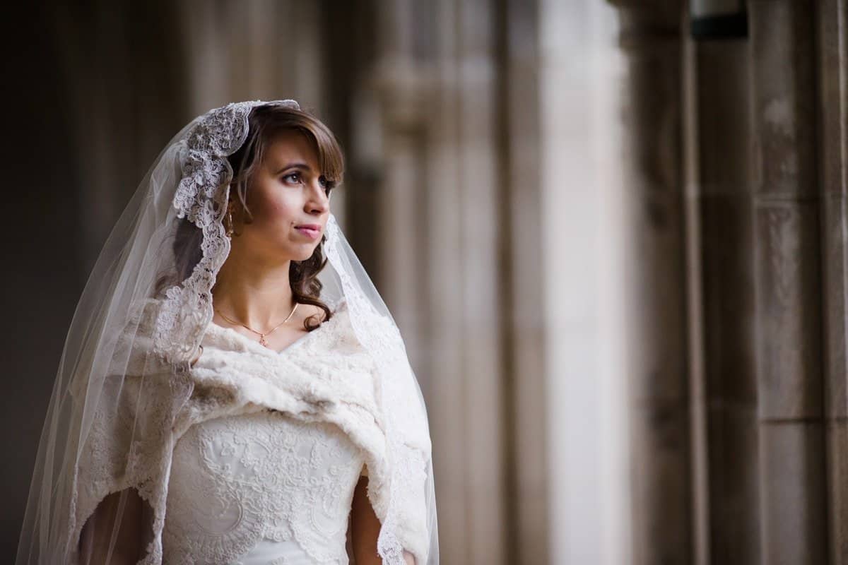 A bride in a white lace wedding dress and veil stands in a softly lit corridor, gazing out a window. The setting is an elegant, arched hallway with stone columns. Her expression is serene and contemplative.