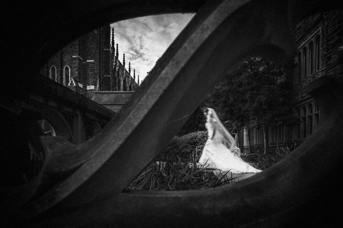 A black and white photo of a bride in a flowing gown walking past a gothic-style building. The foreground features an archway framing the scene, providing a dramatic, artistic perspective.