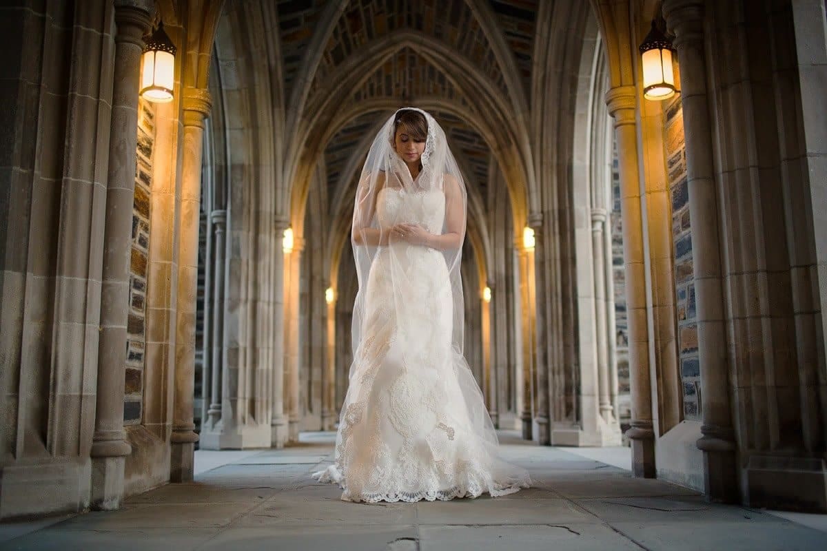 A bride in a white gown stands under a series of gothic arches, softly lit by wall lamps. She is looking downward, with her hands gently clasped. The scene is serene and elegant, with an architectural background.