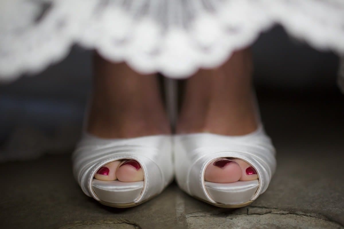 A close-up of a brides feet wearing white, open-toe satin heels. Her toenails are painted a deep red, and part of her white lace dress is visible above. The focus highlights the elegance and detail of the shoes and pedicure.