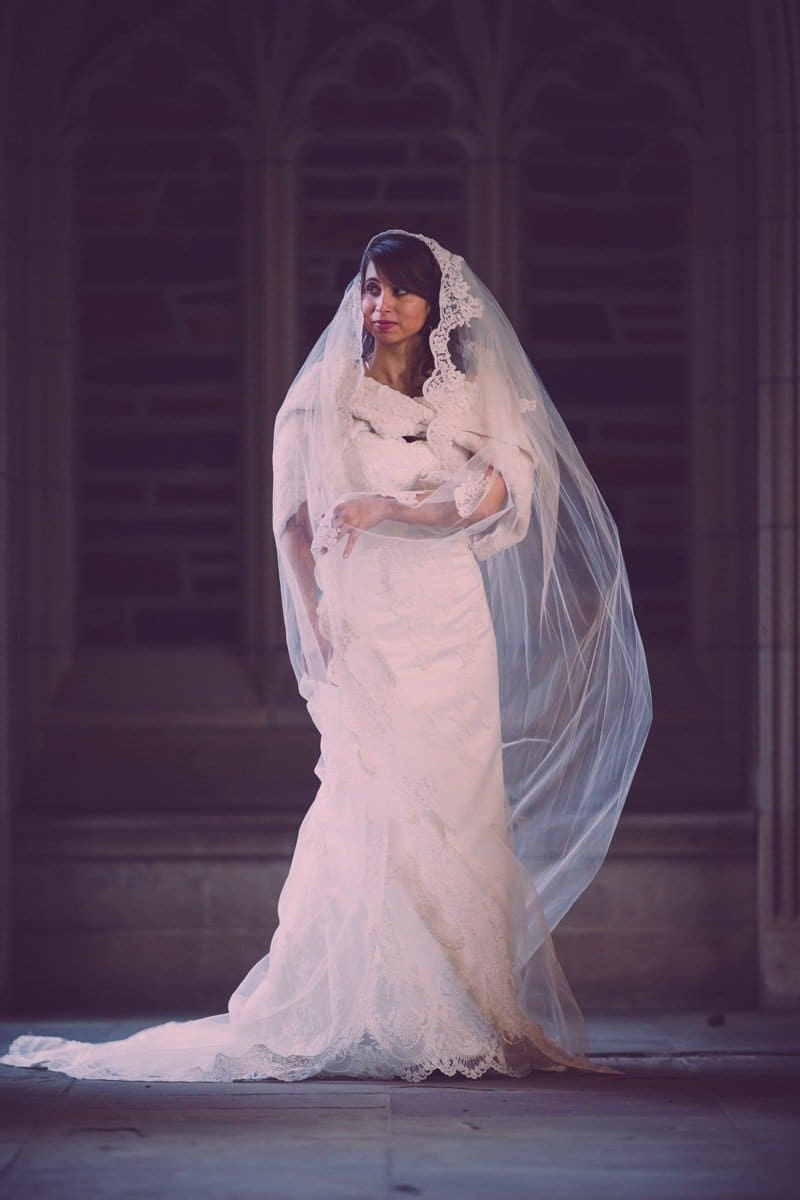 Bride in a white lace dress with a long veil stands in an elegant pose. She is in a shadowed, arched setting with a dim, dramatic light casting a serene ambiance.