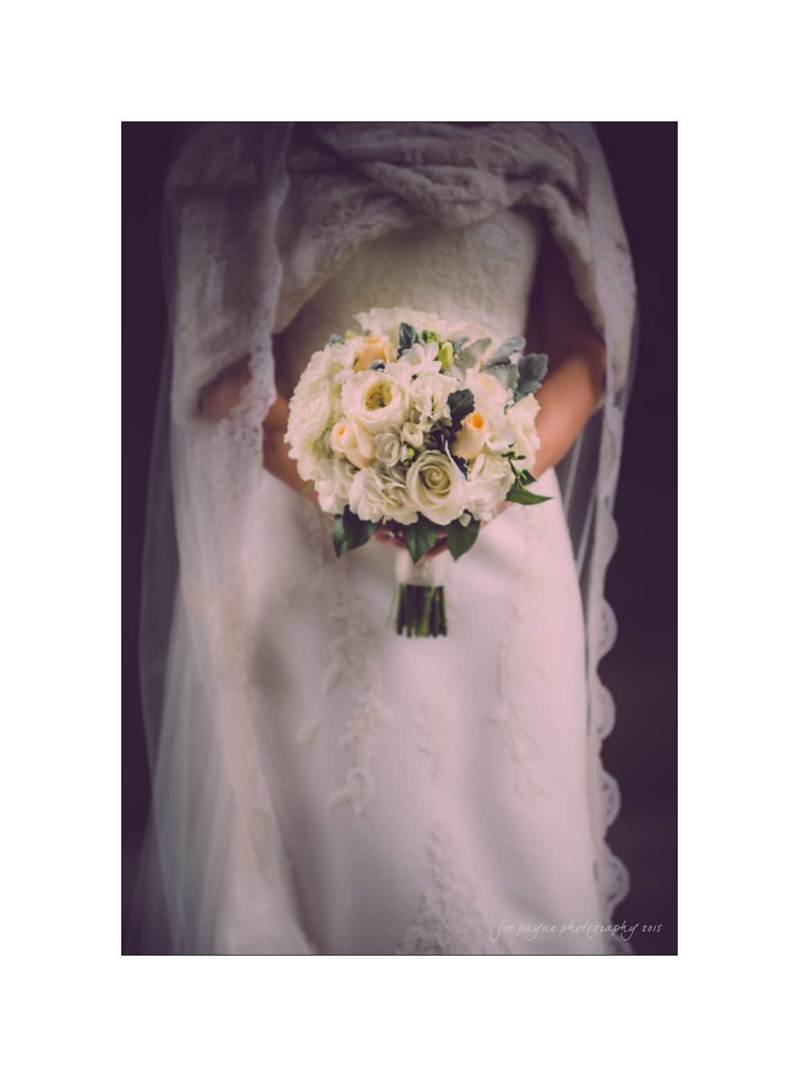 A bride in a lace wedding dress holds a bouquet of white and yellow flowers. Her veil drapes over her shoulders, creating an elegant silhouette. The background is softly blurred, highlighting the bouquets delicate details.