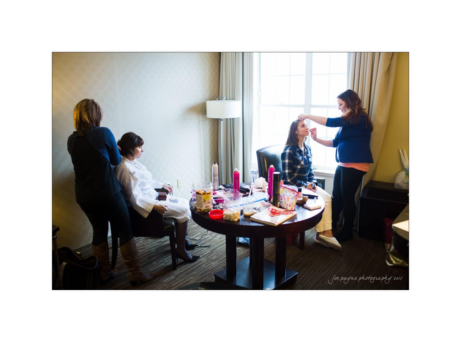 Two women are preparing for an event in a bright room. One is seated, getting her hair styled, while another is having her makeup done. Stylists are focused on their work. A large window with natural light is in the background.
