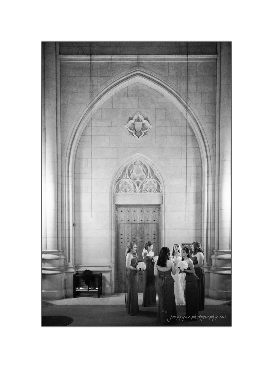 A group of women in formal dresses stand in a semi-circle under a large, ornate stone archway with a wooden door. The setting appears to be inside a grand building, like a church or cathedral. The image is in black and white.