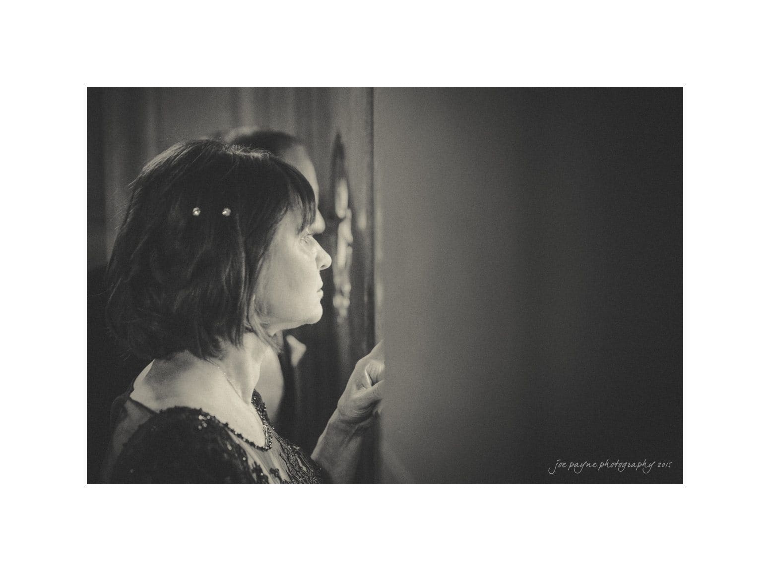 A woman with dark hair and small hairpins, dressed in a dark top, gazes intently at something off-frame. The scene is in black and white, creating a moody atmosphere, with another partially obscured person in the background.