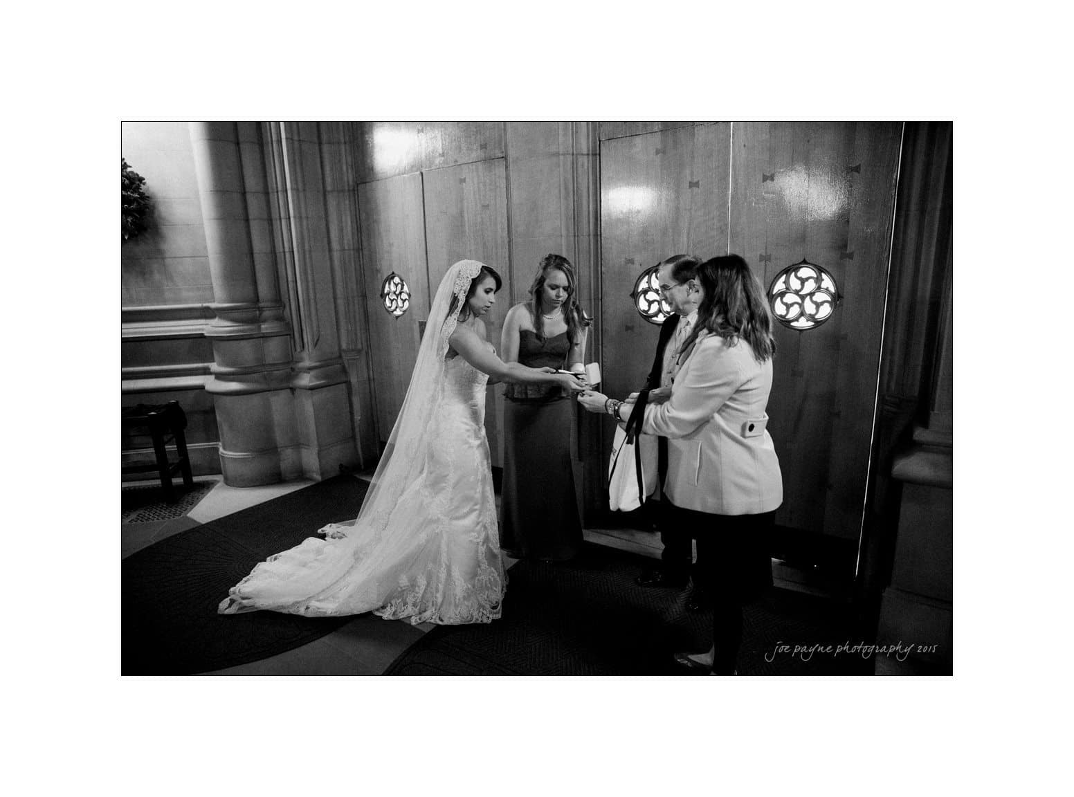 A black and white photo of a bride in a wedding dress holding hands with a woman in a light coat. Two other people stand nearby, including one in a dark dress. They are standing inside a building with intricate wall details.