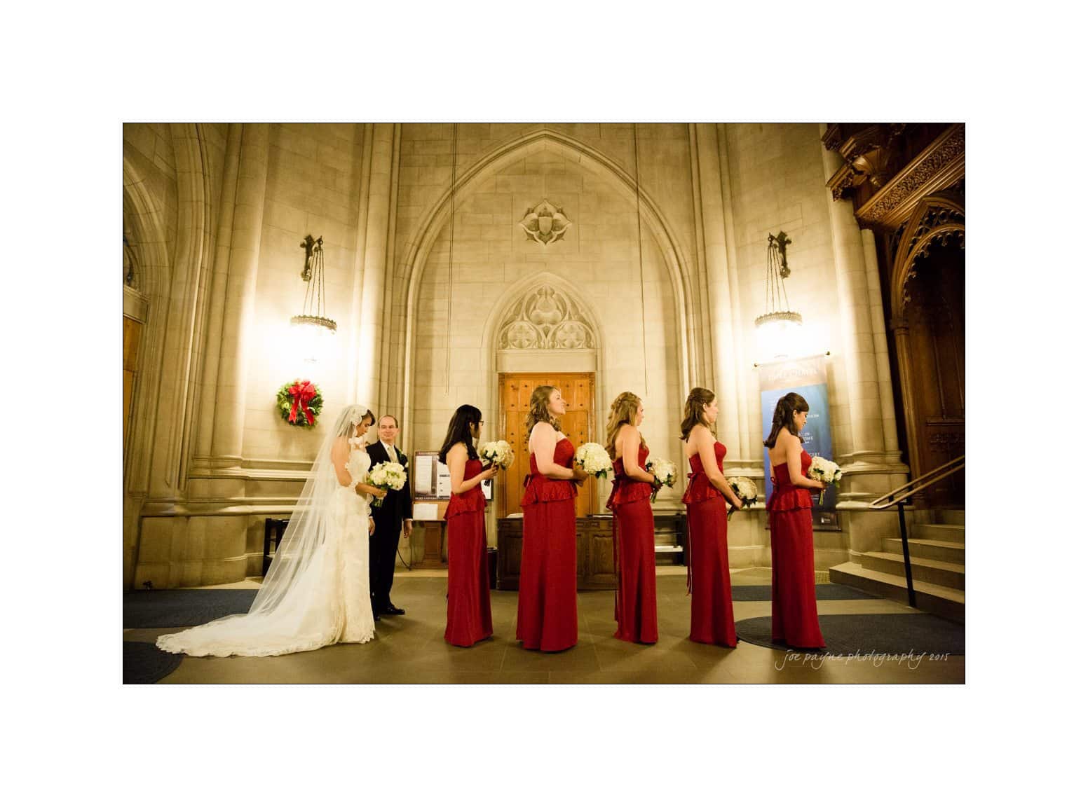 A bride in a white gown and veil stands with five bridesmaids in red dresses, holding bouquets. They are in a church with high stone walls, ornate wooden doors, and warm lighting. A holiday wreath hangs on the wall.