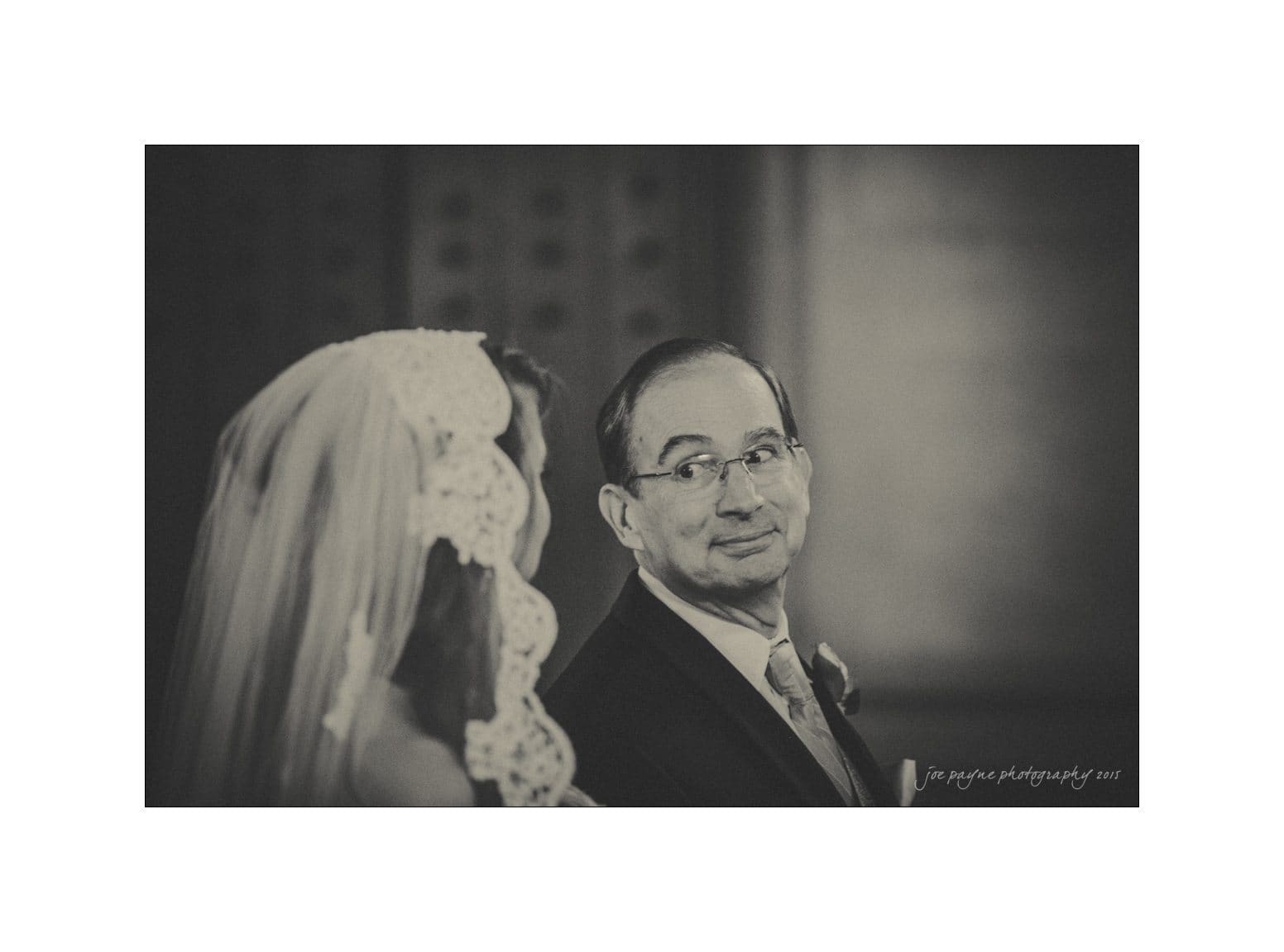 A black and white photo shows a bride in a veil looking at a man in glasses and a suit, who is warmly smiling back at her. The image has a soft focus and an intimate, candid feel.