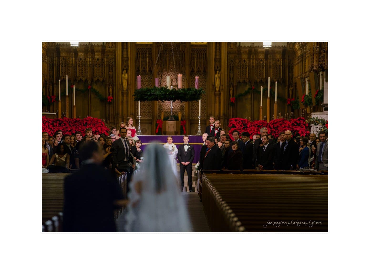 A bride in a white dress walks down the aisle of a large, ornate church filled with people. The groom waits at the altar. The church is decorated with wreaths, poinsettias, and lit candles.