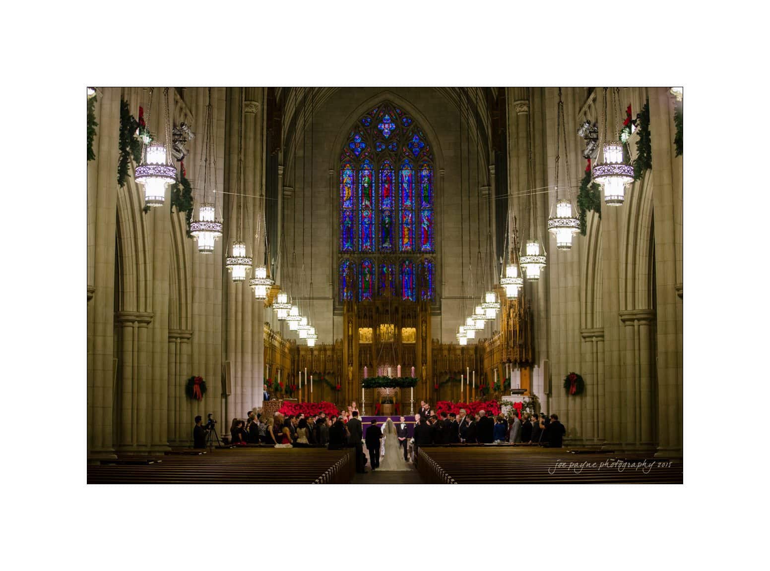 A grand, illuminated church interior with a high arched ceiling and stained glass windows. A large congregation fills the pews, focusing on the altar adorned with red decorations. Hanging chandeliers line the pathway.