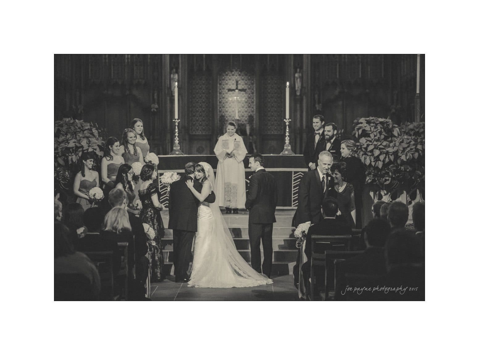 A black and white image of a wedding ceremony in a church. A couple is kissing at the altar as bridesmaids and groomsmen stand nearby. A priest is in the background, with guests seated in pews.