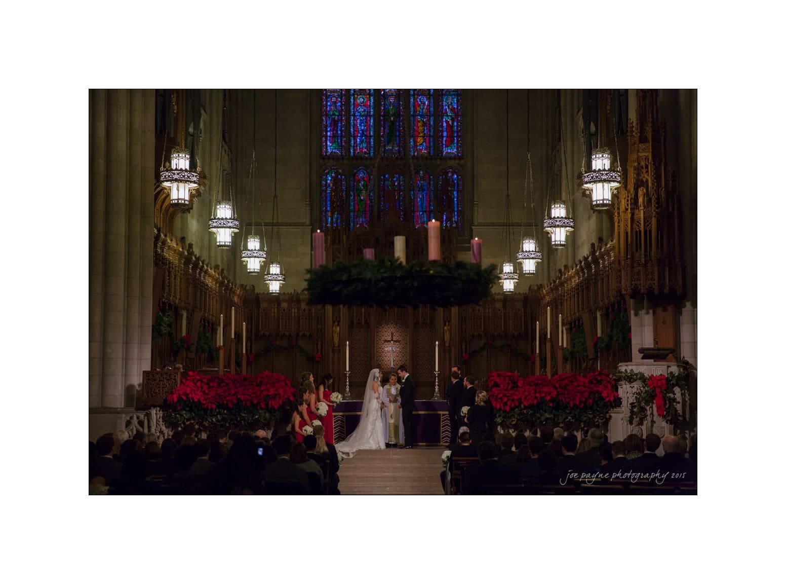 A wedding ceremony in a grand church with a stained glass backdrop. The bride and groom stand at the altar, surrounded by bridesmaids, groomsmen, and red poinsettia decorations. The dim lighting creates a warm, intimate atmosphere.