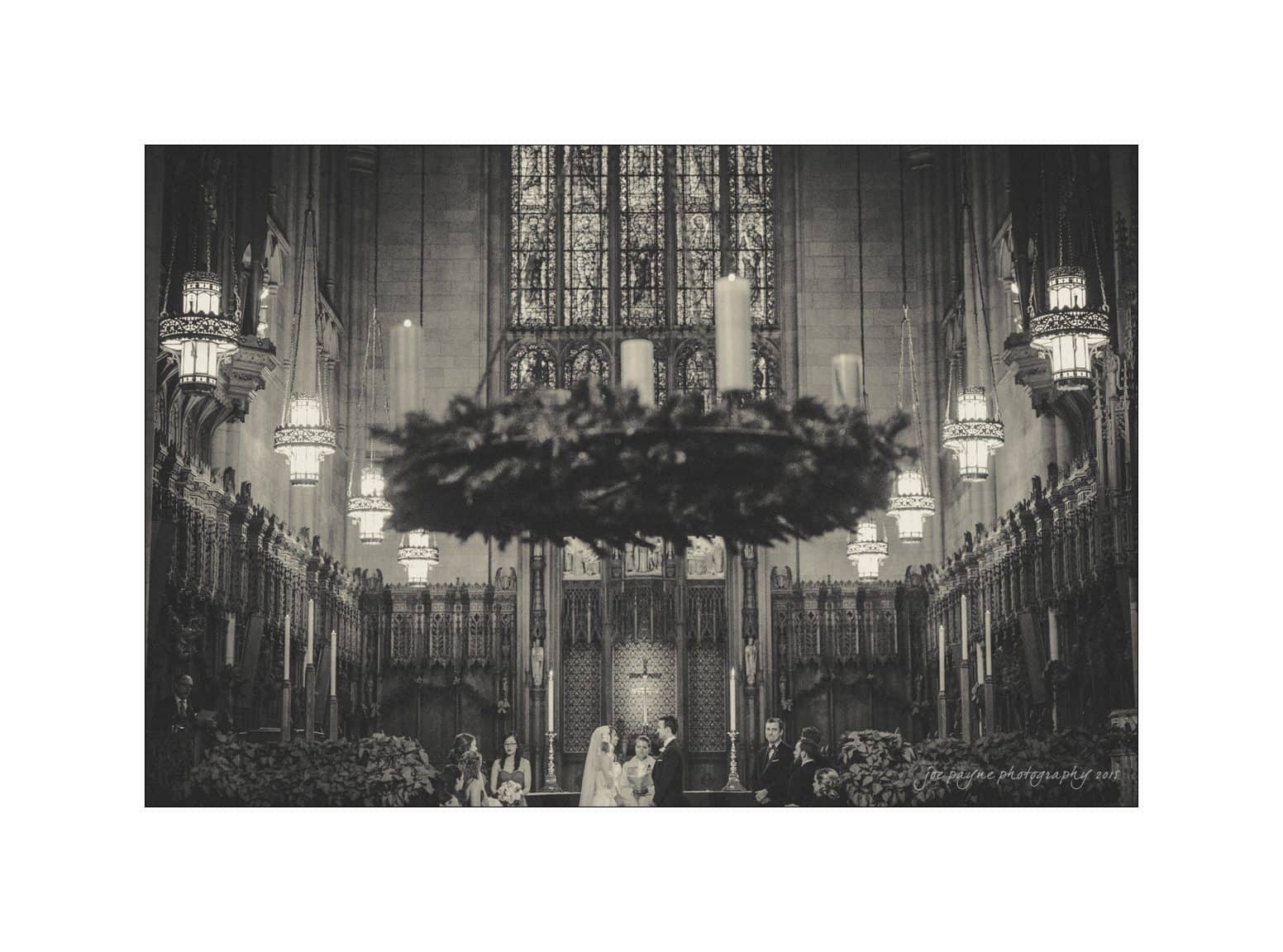 A black-and-white photo of a church interior during a wedding ceremony. A vaulted ceiling, stained glass windows, and ornate chandeliers decorate the space. A large candle wreath hangs in the foreground, slightly obscuring the altar.