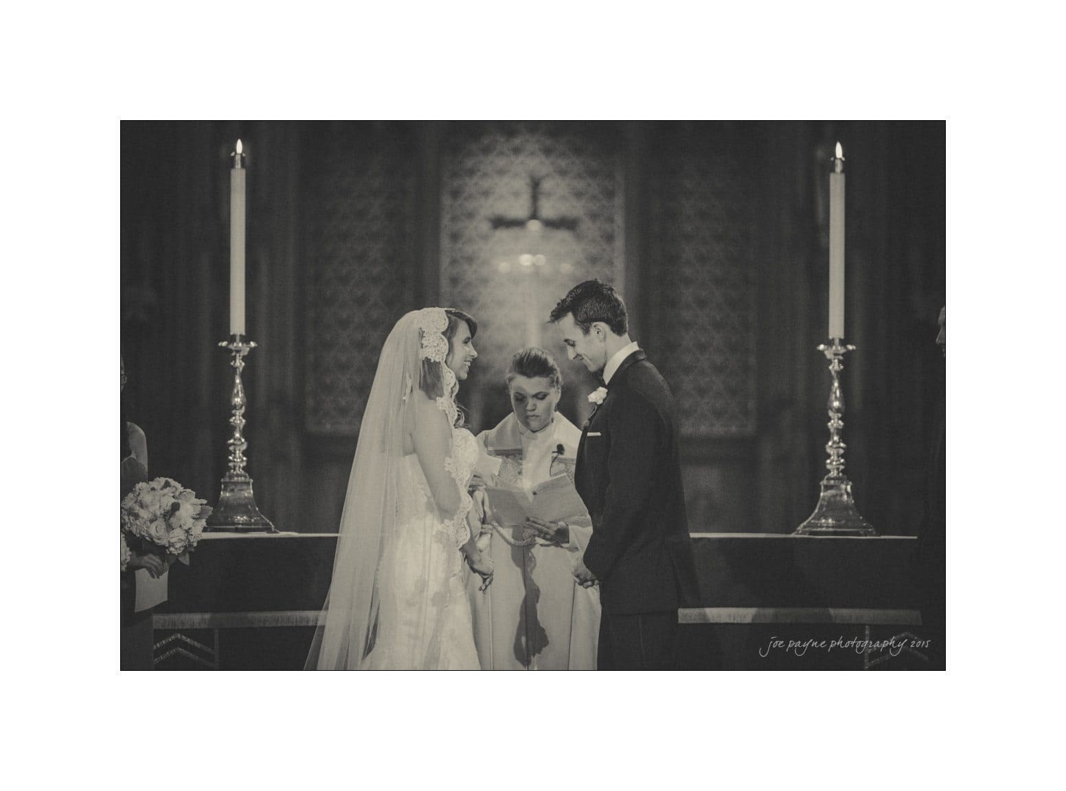 A bride and groom stand at the altar with a minister during a wedding ceremony. The bride is in a gown and veil, and the groom is in a tuxedo. Two tall candles are on either side, and an ornate background is visible. The scene has a vintage tone.