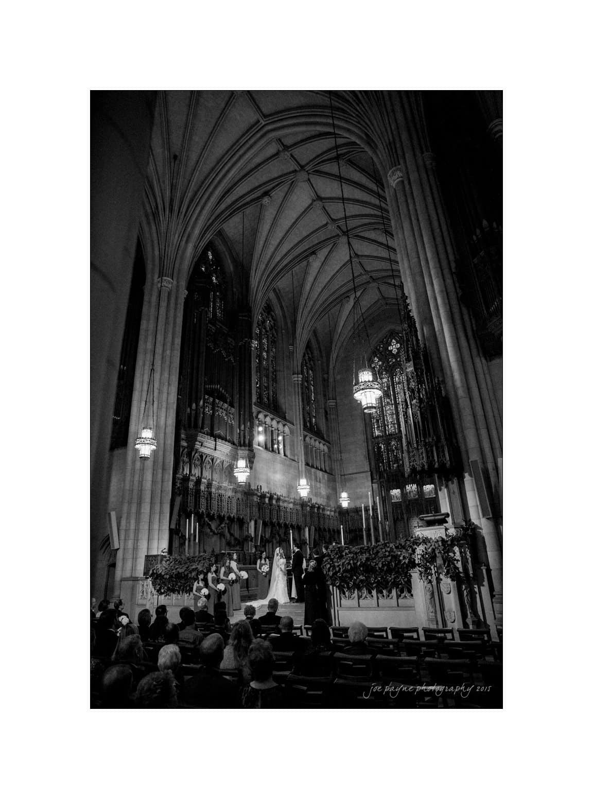 A black and white photo of a spacious cathedral interior with high arched ceilings. A wedding ceremony is taking place at the altar, surrounded by guests. Ornate chandeliers and large windows add to the grandeur of the scene.