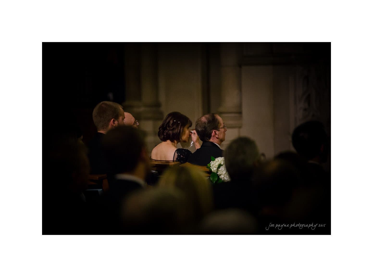 A dimly lit wedding ceremony with an audience seated and attentively watching. A couple is central in the frame, with the woman in an off-shoulder dress and the man in a dark suit. The background features soft architectural details.