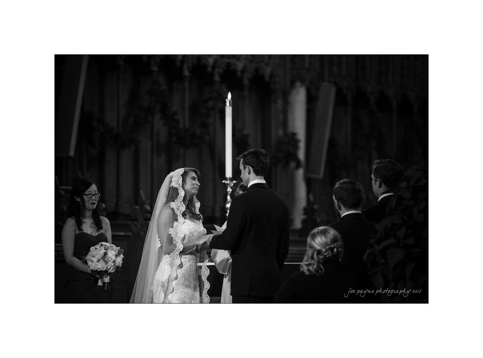 A bride and groom exchange vows in a dimly lit church, with a bridesmaid holding a bouquet nearby. The bride wears a lace veil and gazes lovingly at the groom. A lit candle is in the background.