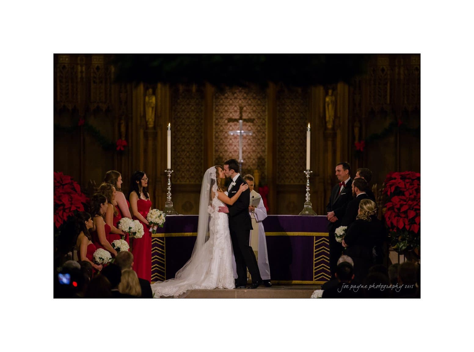 A bride and groom share a kiss at the altar during a wedding ceremony. The bridal party, dressed in red and holding white flowers, stands on either side. The backdrop features a cross, large candles, and festive decorations.