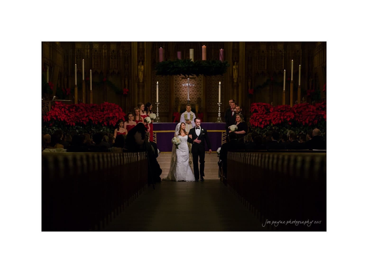 A bride and groom walk down the aisle in a decorated church. Bridesmaids in red dresses stand to the left, with a choir in the background. The scene is illuminated by candles and festive red poinsettia arrangements.