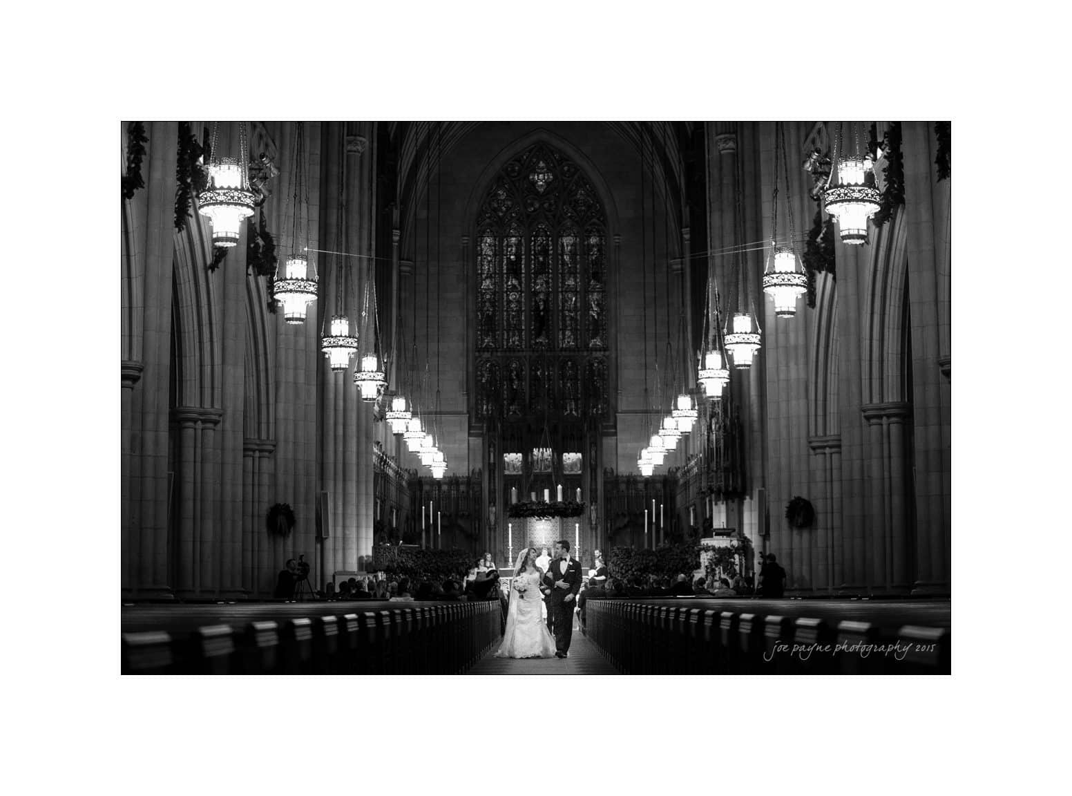 A black and white photo of a couple walking down the aisle of a large, ornate church. The interior features high vaulted ceilings, intricate stained glass, and hanging lamps. The couple is in wedding attire, holding hands.