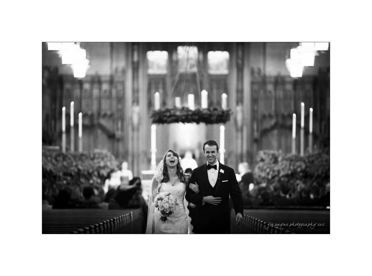 A bride and groom smiling and walking arm in arm down the aisle of a grand church, decorated with chandeliers and floral arrangements. The scene is in black and white, capturing a joyful moment.