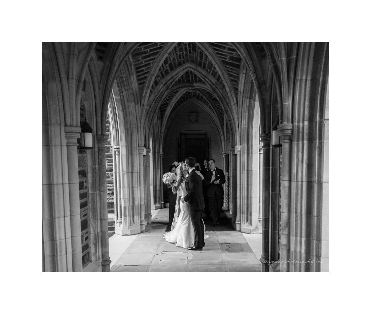 A black and white photo of a wedding couple kissing in a stone archway, with a best man and a woman standing behind them. The setting is a cathedral-like structure with intricate arches and light coming from the left.