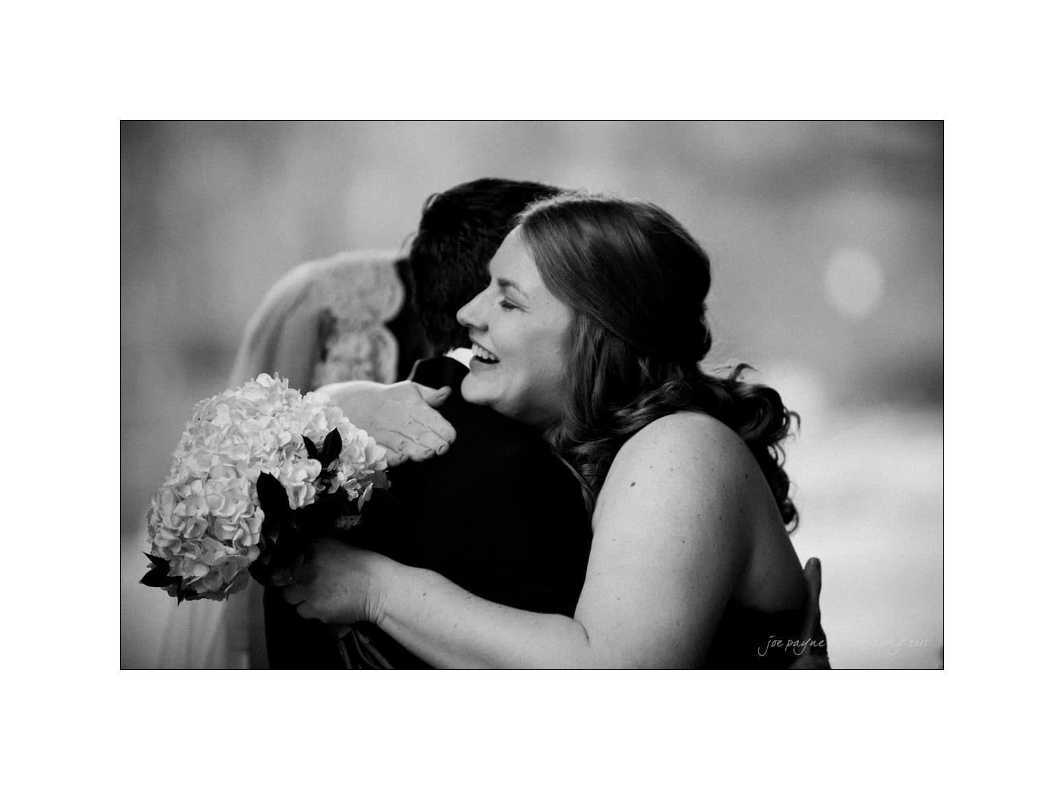 A black and white photo shows a smiling woman with long hair embracing another person who is partially visible. She holds a bouquet of flowers in her left hand, capturing a moment of joy and affection.