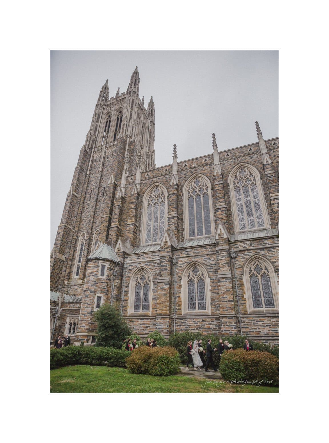 A large, gothic-style cathedral with tall stained-glass windows and a towering spire. People walk nearby on a cloudy day, surrounded by greenery.